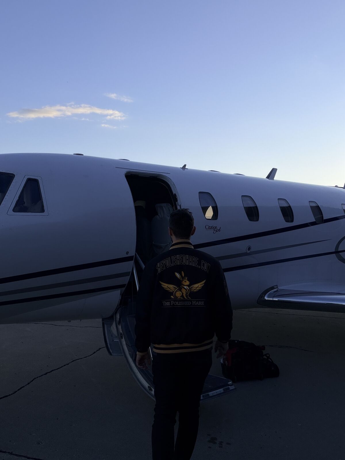The Polished Hare aviation detailer boarding Cessna Citation Excel wearing branded jacket with equipment bag on ramp at dusk