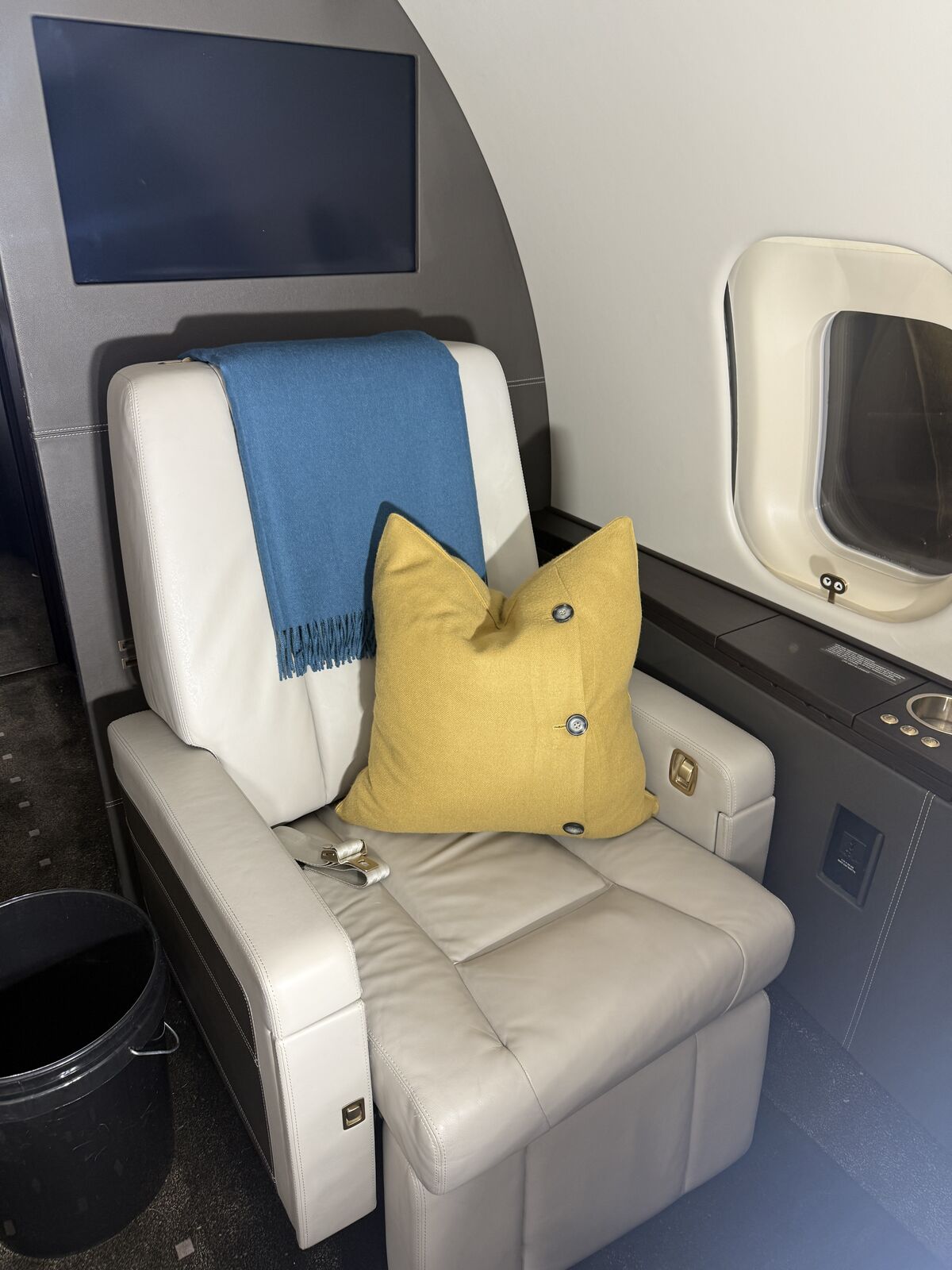 Private jet white leather passenger seat with yellow accent pillow and blue throw blanket beside cabin window after interior detailing